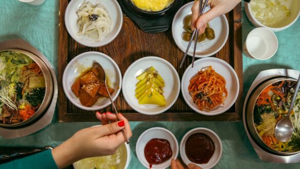 An aerial view of a table filled with bowls of various kinds of food and hands with chopsticks coming into frame to grab a bite.