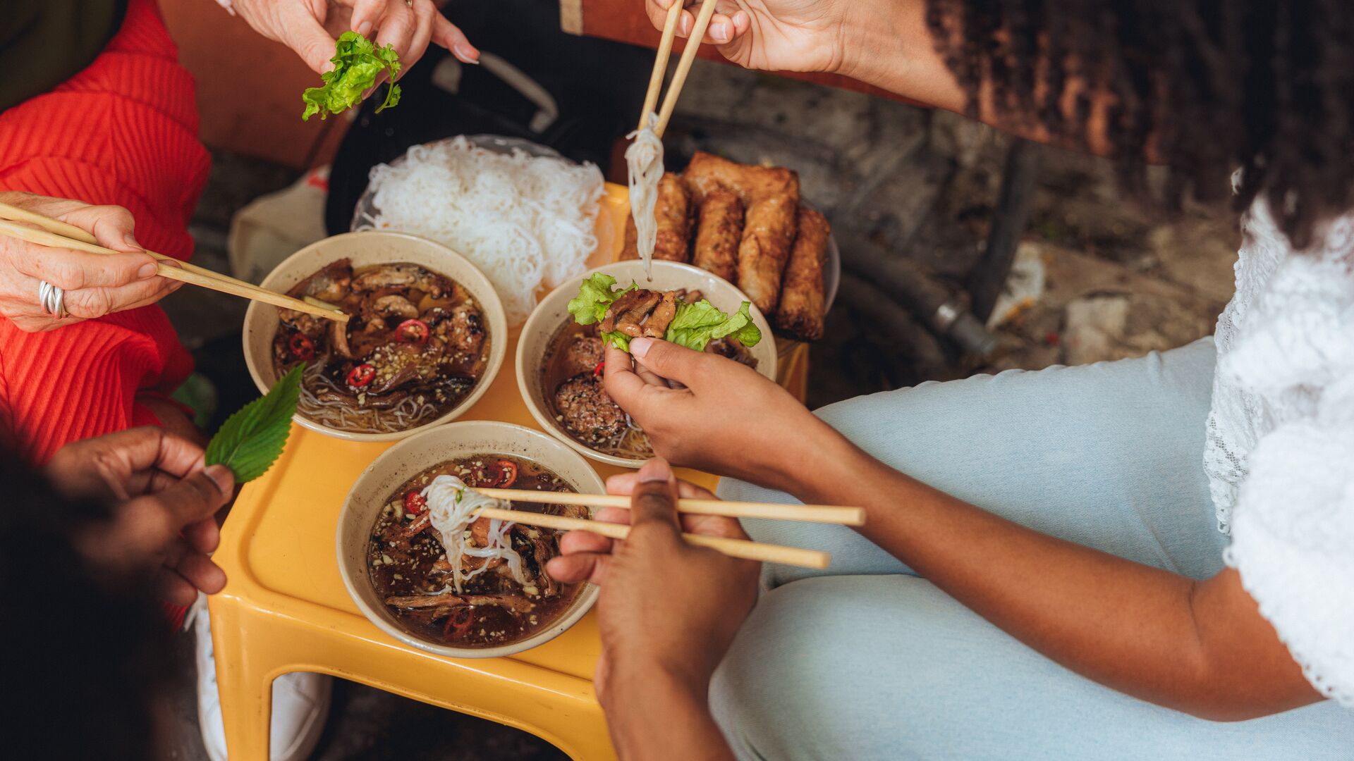 Three travellers sit at a low table in Vietnam eating a meat and noodle dish with chopsticks. 
