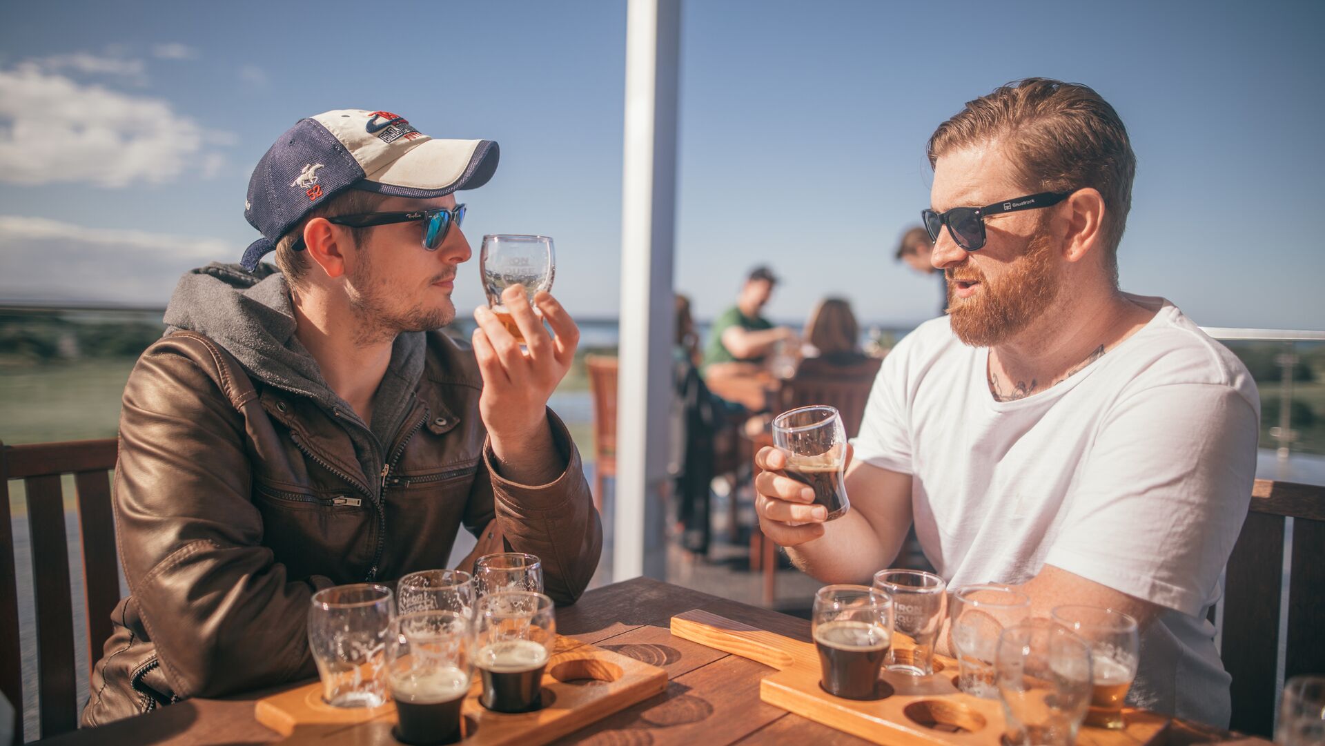 Two men drink from small beer glasses with a paddle of beer on the table in front of them.