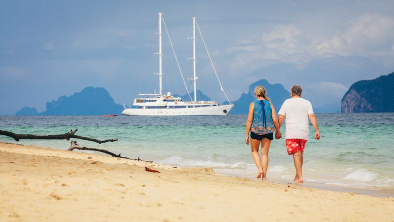 A couple walking down the beach in Thailand with an Adventure Cruise ship in the sea in front of them.