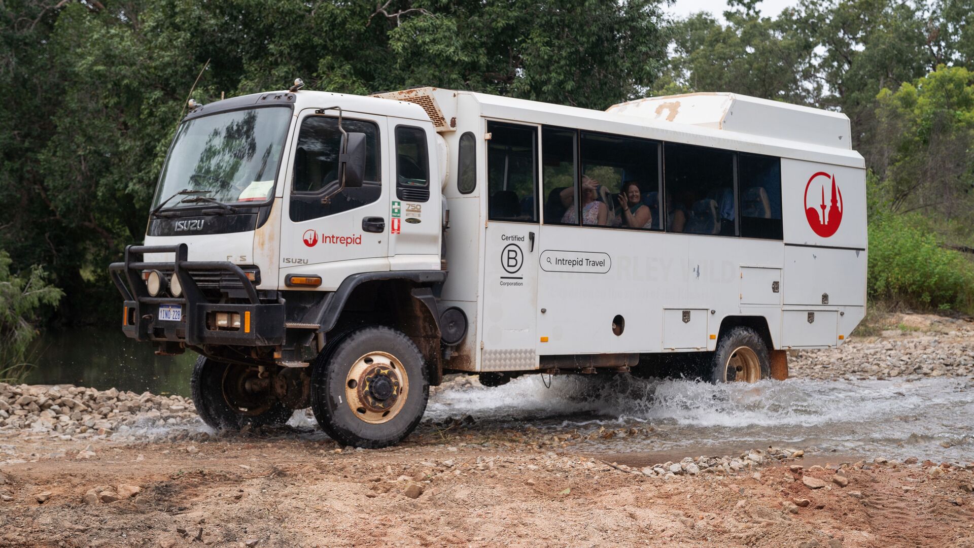 An Intrepid Overland truck crosses a creek with passengers smiling inside.