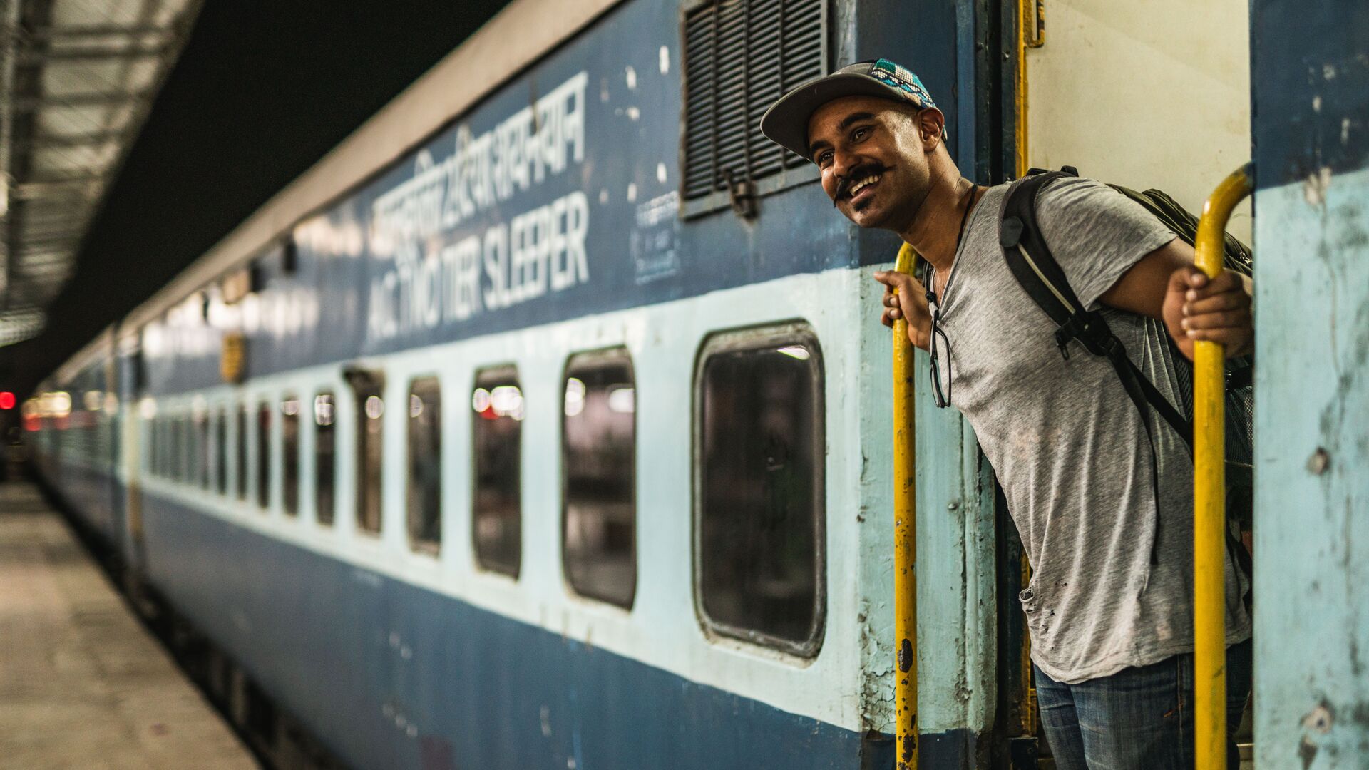A man stands at the door of a train in India smiling.