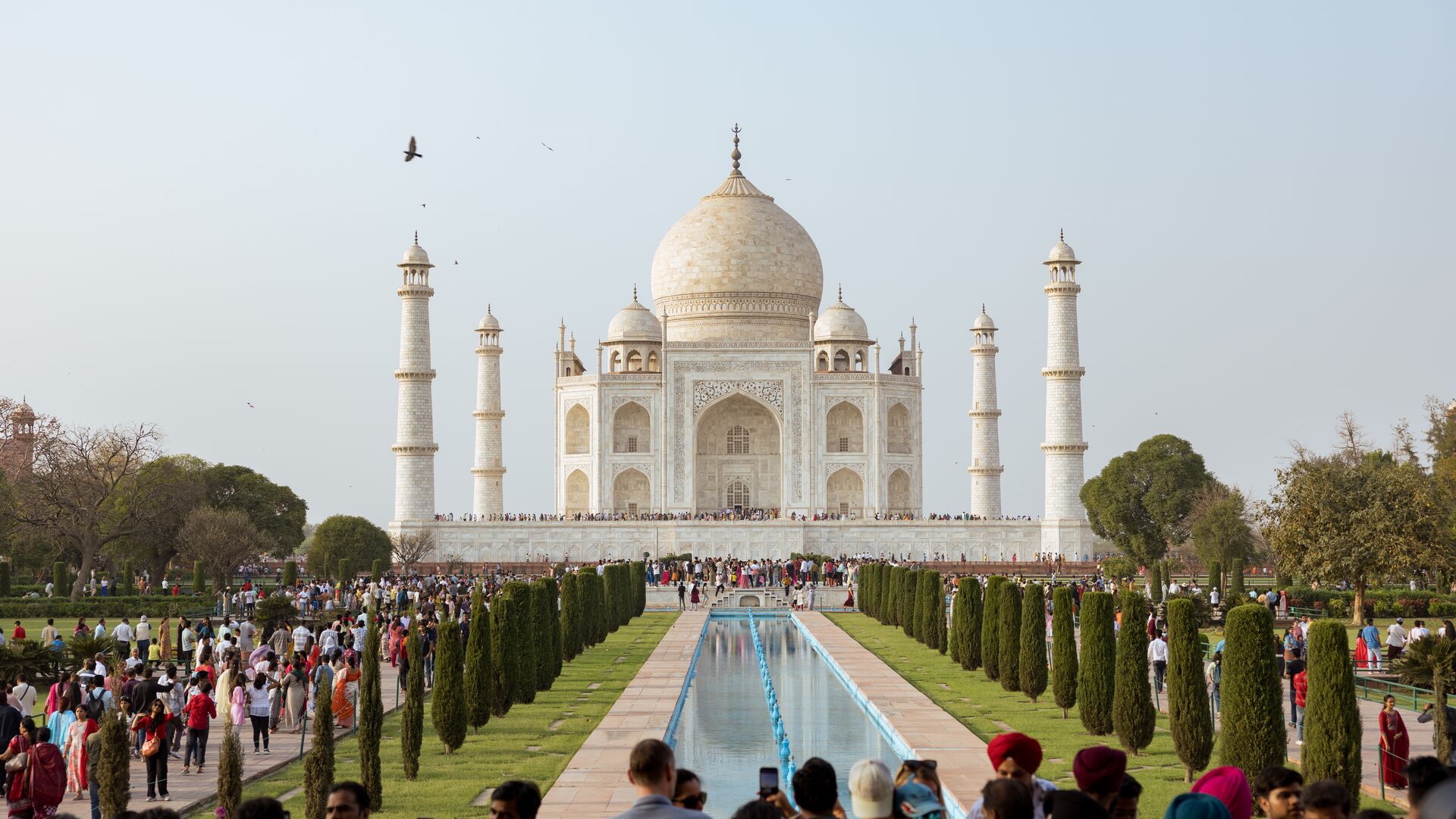 The Taj Mahal made out of white marble in the background with the reflecting pool coming down through the middle of the photo and crowds of people on either side of the long, rectangle reflection pool. 