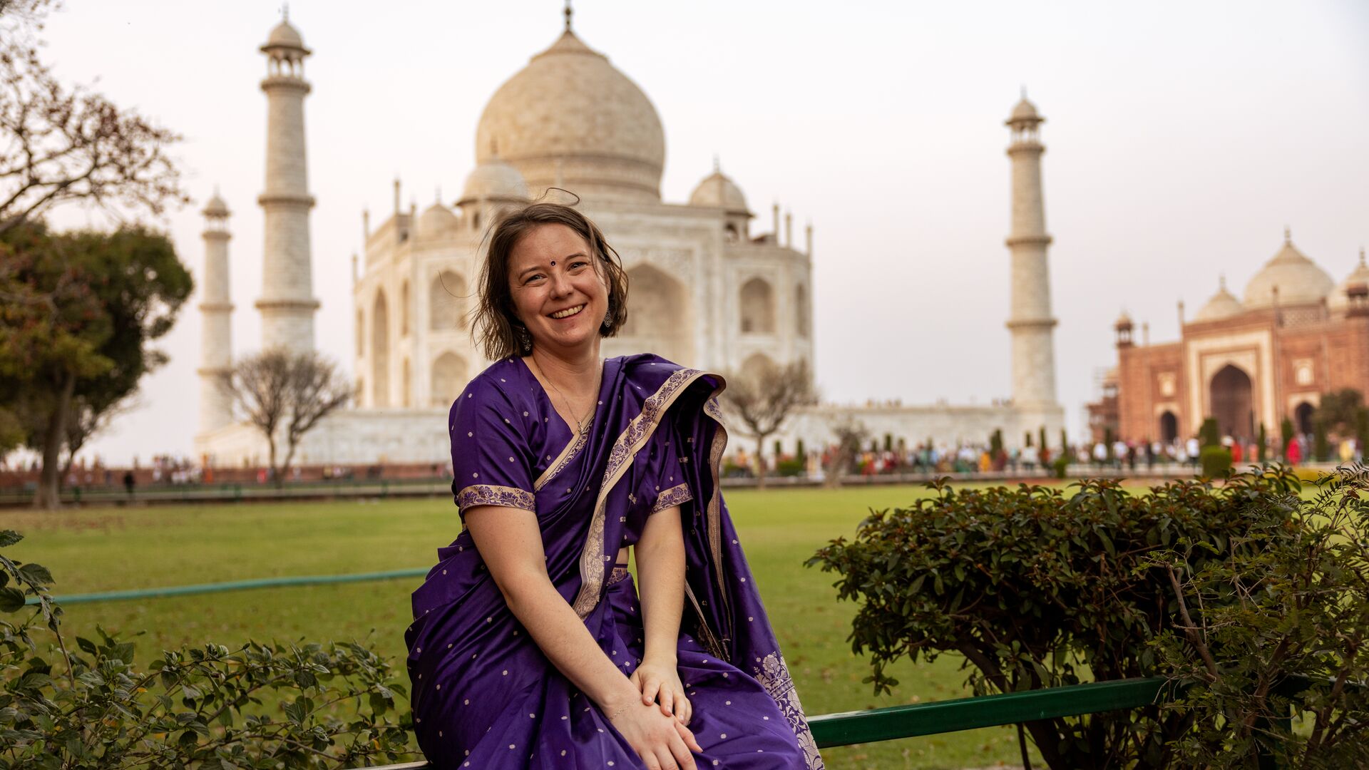 A woman in a purple saree poses for a photo in front of the Taj Mahal. 