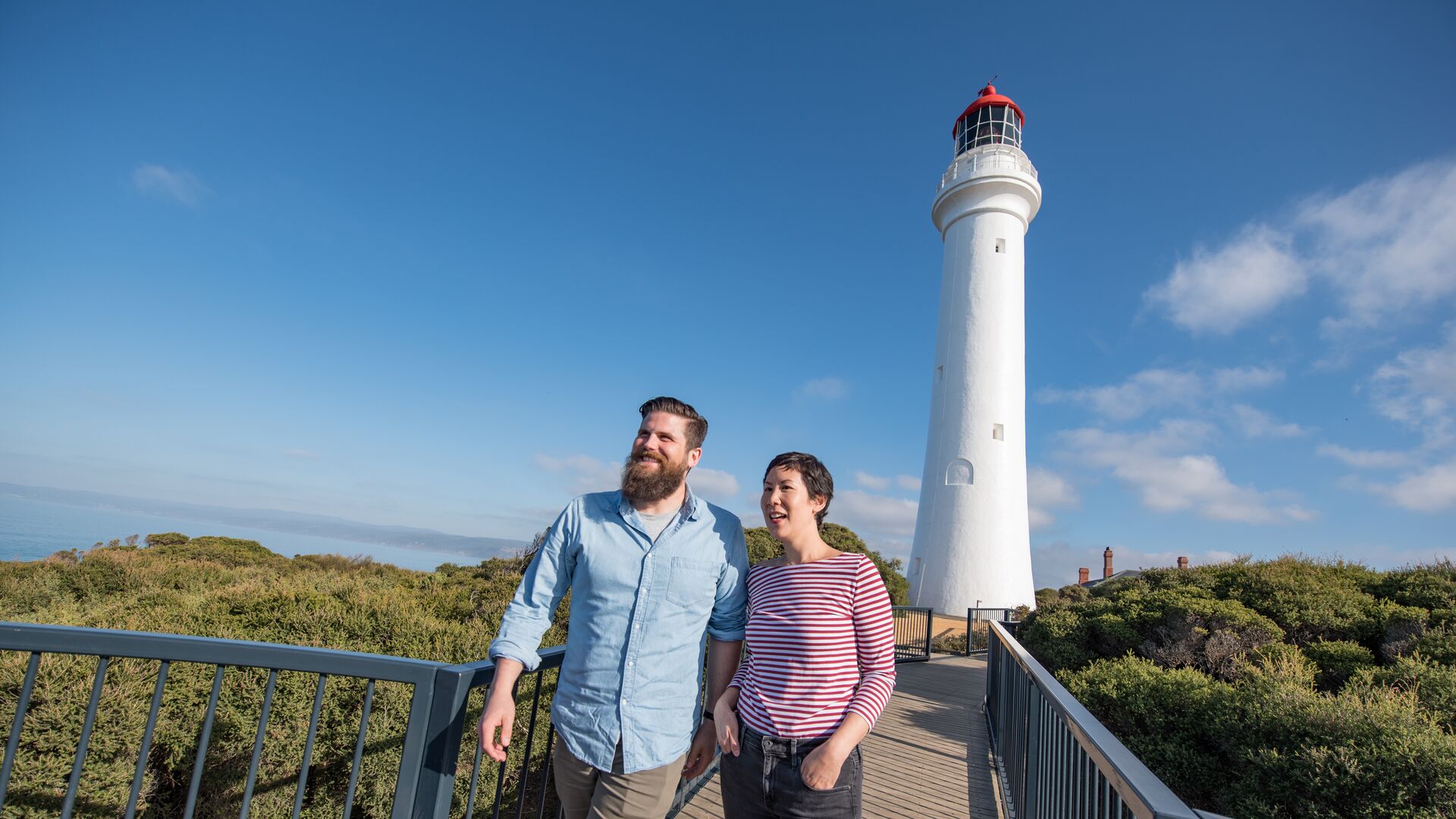 Two people walk along a boardwalk down from the Aireys Inlet Lighthouse.