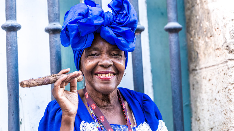 Portrait of African Cuban woman with blue headdress and clothing, happily smoking a cigar in Havana, Cuba. 