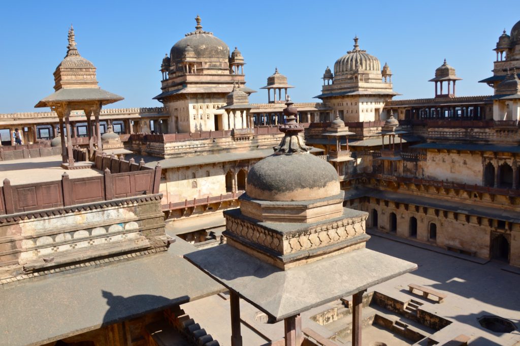 Rooftop view of Chaturbhuj Temple complex in Orchha, showing multiple stone domes, ornate pavilions and galleries in sandstone and dark brown, under a clear blue sky