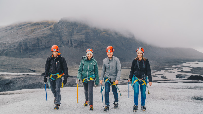 4 smiling travellers dressed in helmets and climbing gear pose in front of a glacier.