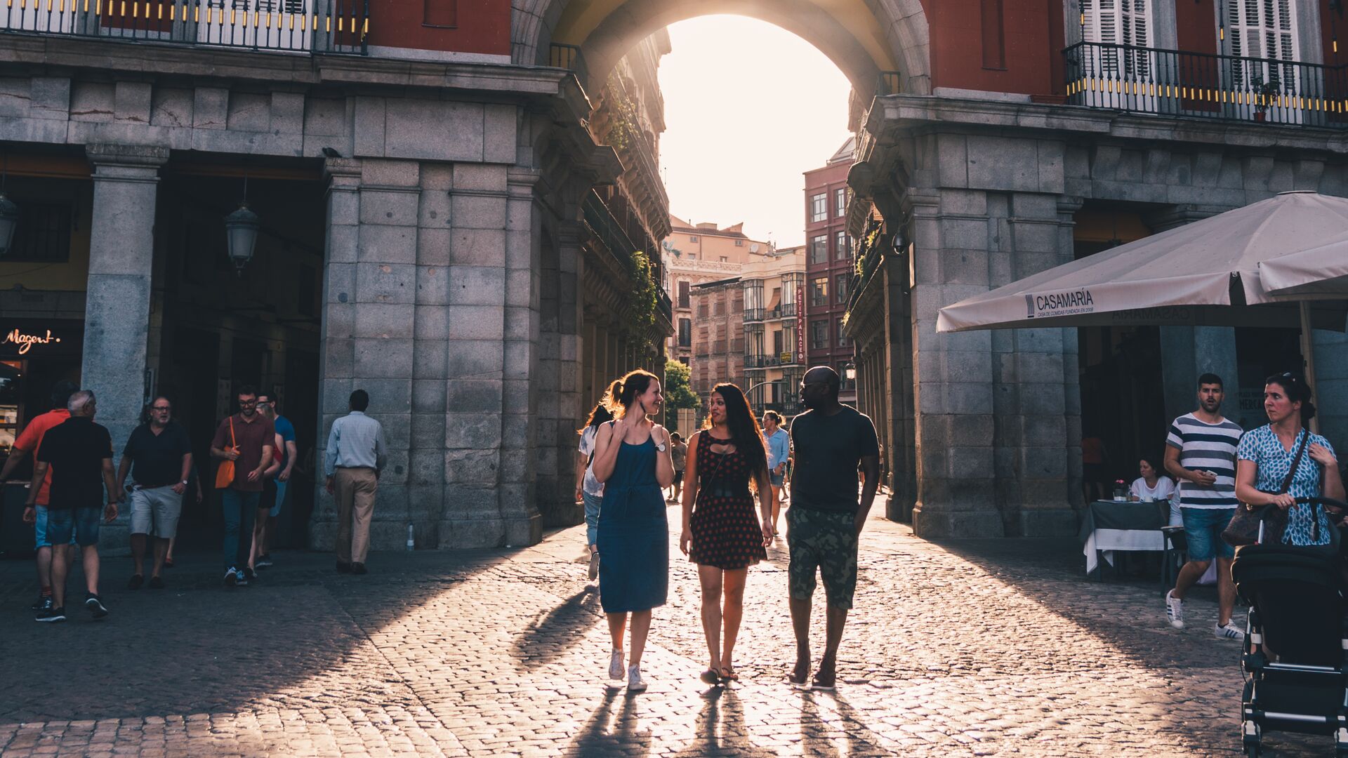 A group of three travellers walking through an arch in a plaza in Madrid, Spain, with the sun casting a path on them while they chat.