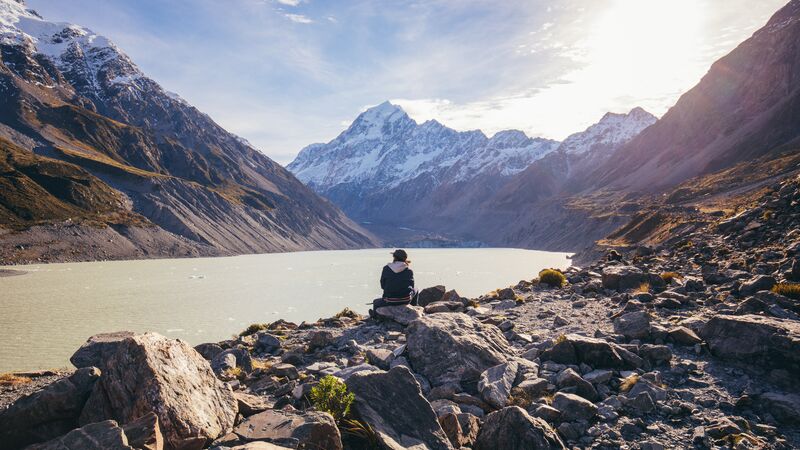 A man sits on a rock in front of a beautiful lake and mountains