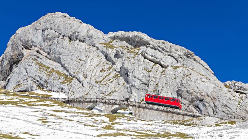 A small red train beside a large stony mountain