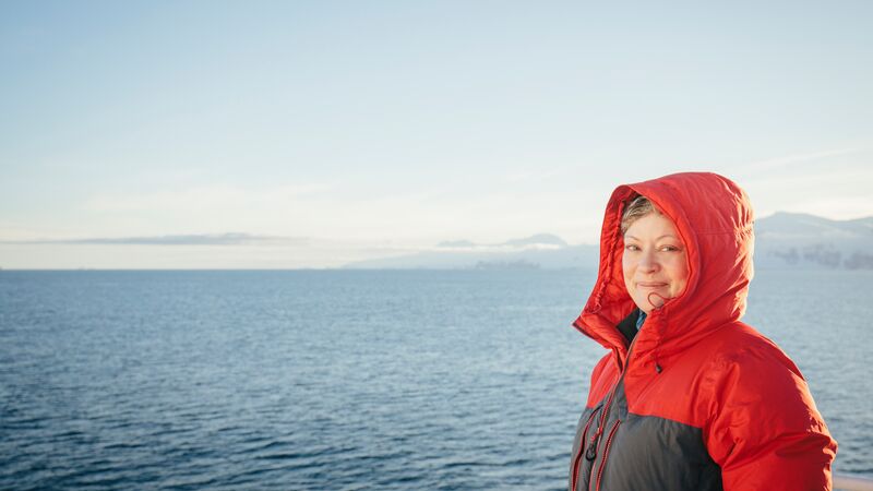 A woman in a red jacket smiling at the camera with the ocean behind her