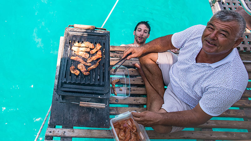 A man cooks at a barbecue on a boat, while another man pops his head out of the water to look