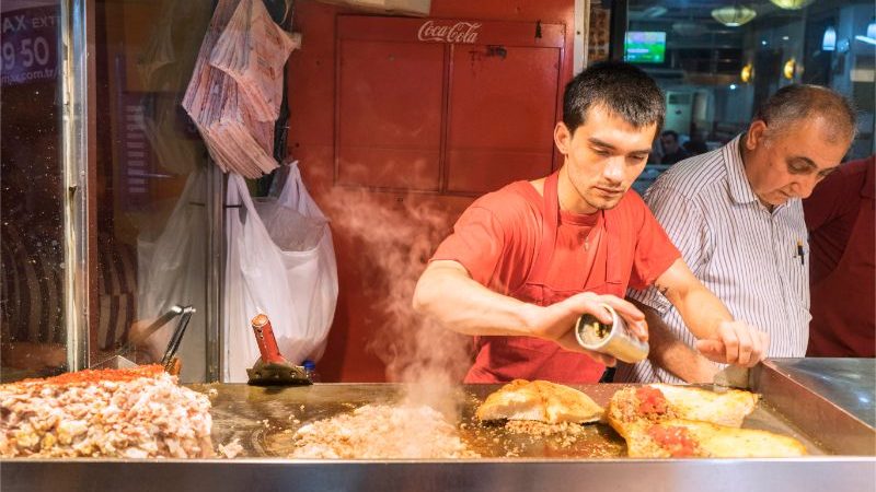 A man in a red t-shirt at a market prepares a snack over a stove