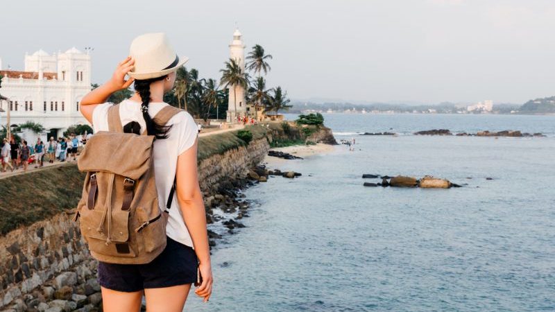 A woman wearing a hat and a backpack admiring the ocean and a lighthouse in Sri Lanka