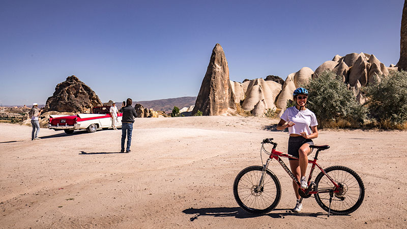 A woman on a bike smiles at the camera with a vintage car in the background