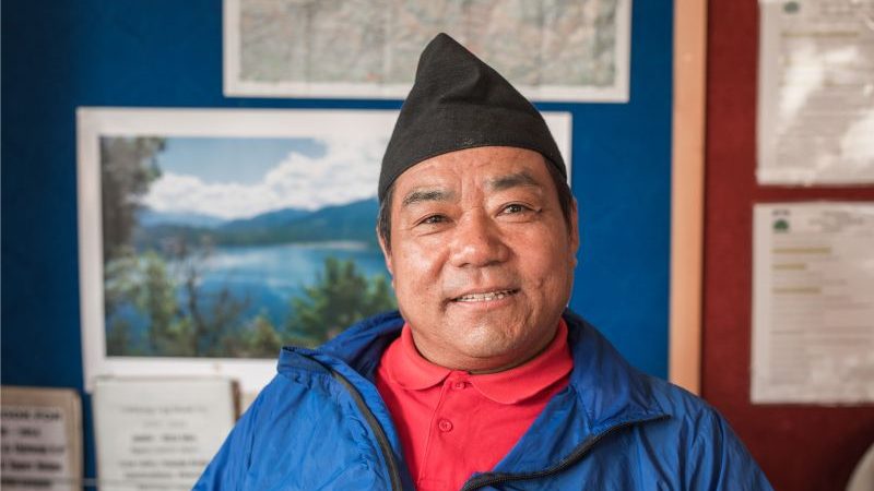 A smiling man wearing a blue top and a black Nepalese hat.