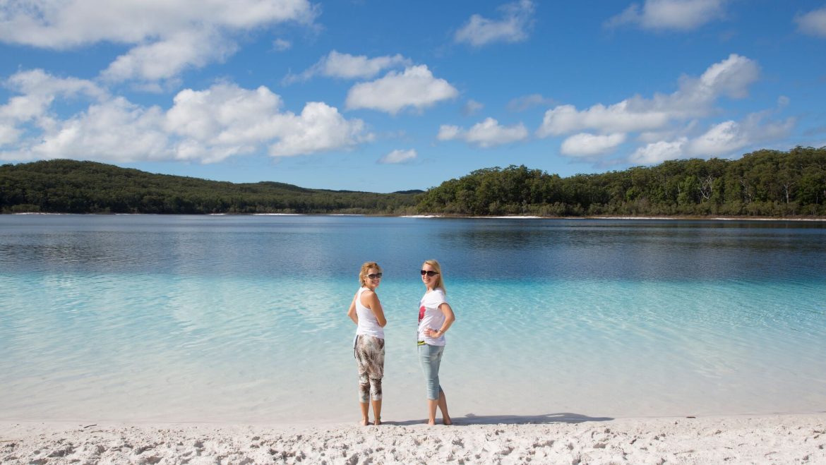 Two women stand on white sand in the clear waters of K'gari