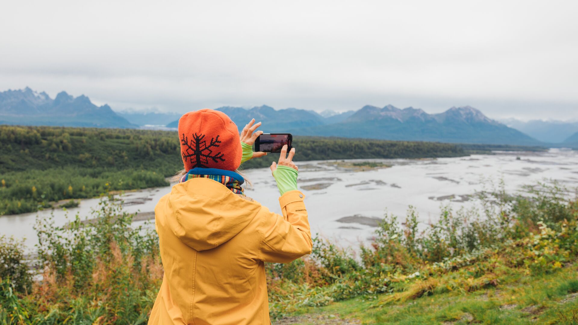 A woman in a yellow rain jacket taking a picture of the mountains in Denali National Park, Alaska.