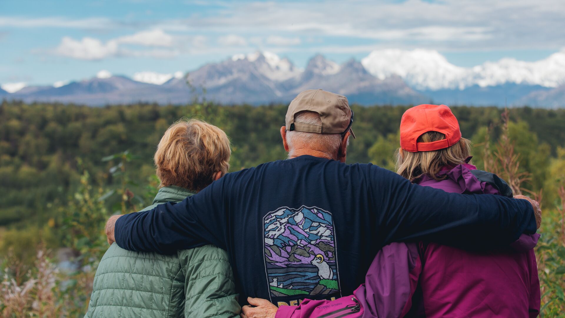 Three travellers stand with their backs to the camera, arms around each other looking out at snow-capped mountains in Denali National Park.