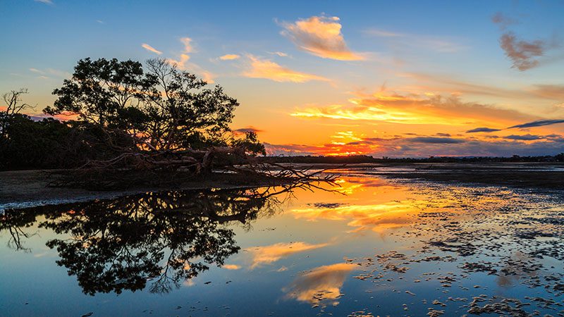 The sun is setting over Nudgee Beach with the colours reflecting on the water.