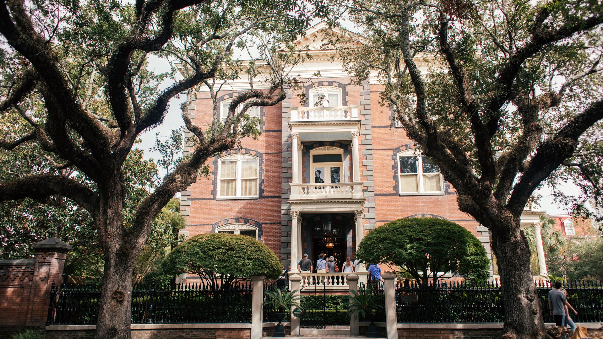 An old brick home in South Caroline with big trees out front and travellers standing at the door. 