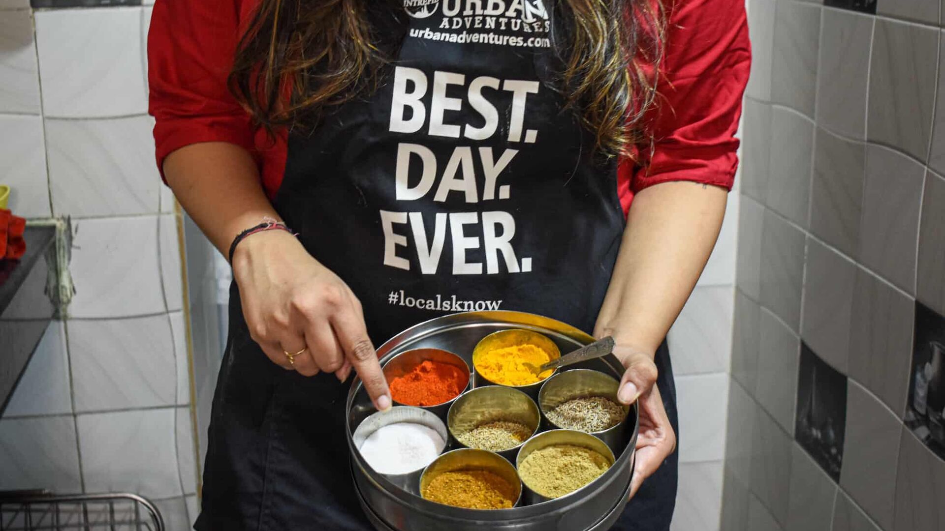A woman in an Intrepid Urban Adventures apron that says 'Best.Day.Ever.' holding and pointing at bowls of spices in a kitchen in India.