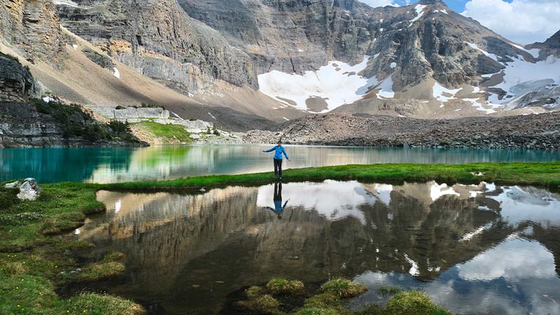 Woman standing in front of mirrored surface of Lake O'Hara with snow-covered mountains in the background.