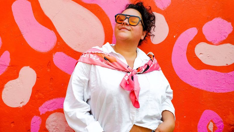 A woman wearing sunglasses and a white shirt stands in front of a pink and red painted wall