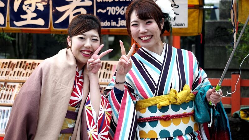Two Geisha women in traditional dress, smiling and holding up the peace sign to the camera. 