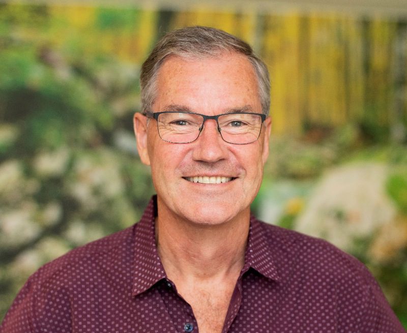 A headshot of a smiling man wearing glasses and a maroon-coloured shirt.