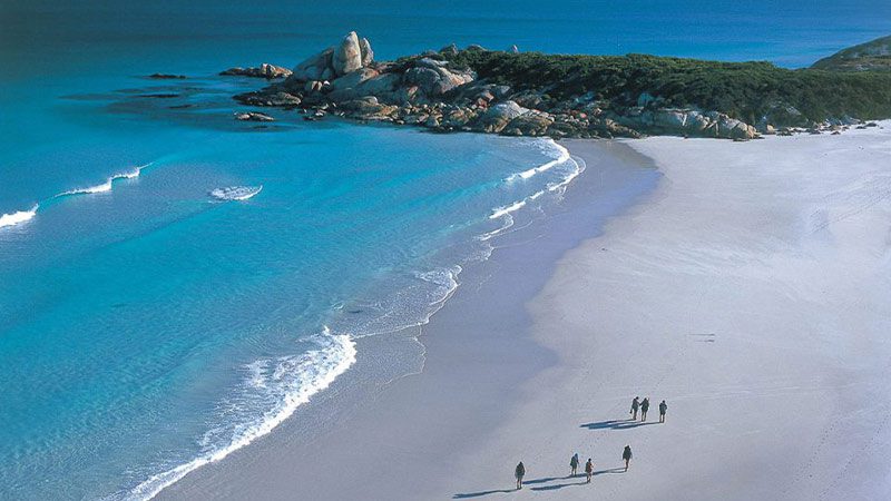 A group of travellers walking along a white sandy beach beside crystal clear water in the Bay of Fires region