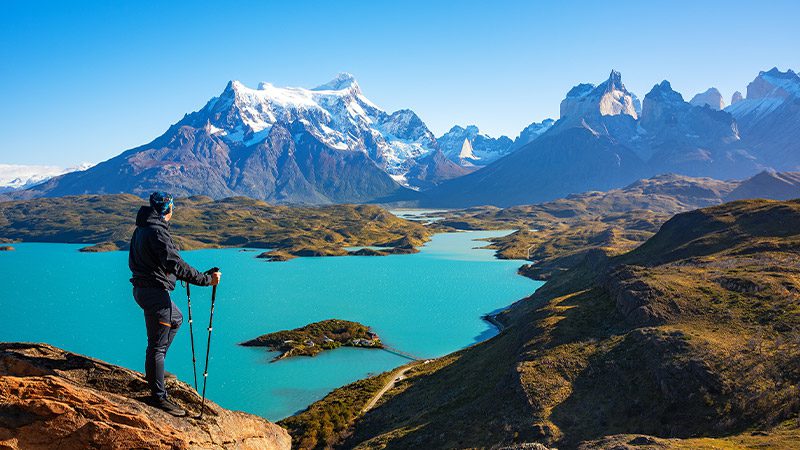 A hiker stands on a mountain overlooking a turquoise lake with snow-covered mountains in the background