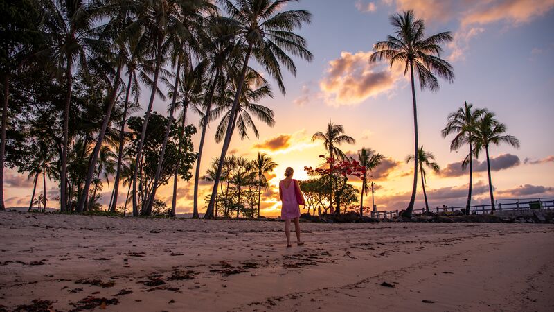 Woman walking along a beach lined with palm trees in Port Douglas at sunset.