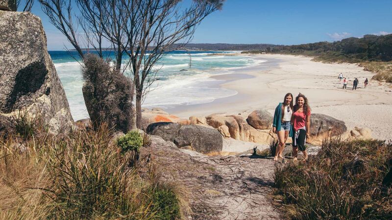 Two travellers smiling at the camera while standing at the sandy entrance to a beach along the Bay of Fires.