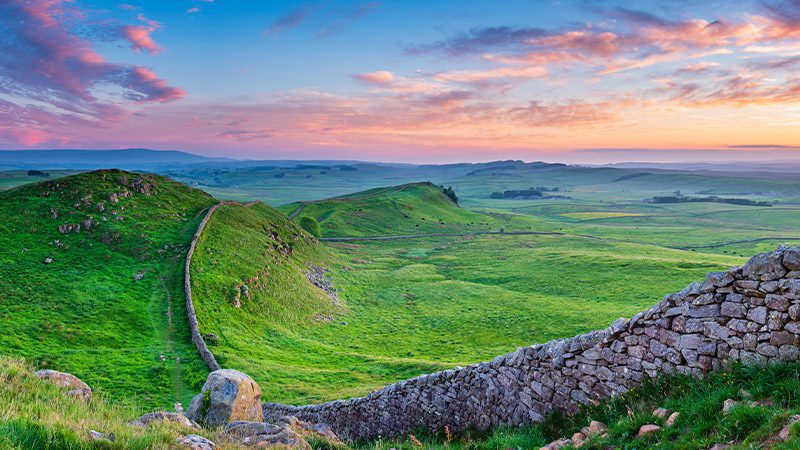 A stretch of Hadrian's Wall in England beside green hills at sunset. 