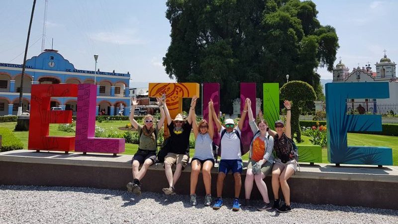 Six happy travellers sitting in front of a colourful sign in Mexico