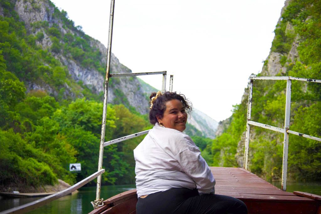 A woman sitting at the front of a boat moving along a river between wooded mountains turns and smiles at the camera
