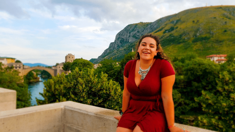 A woman in a red dress sitting on a balcony with a bridge and tree-covered mountains in the background