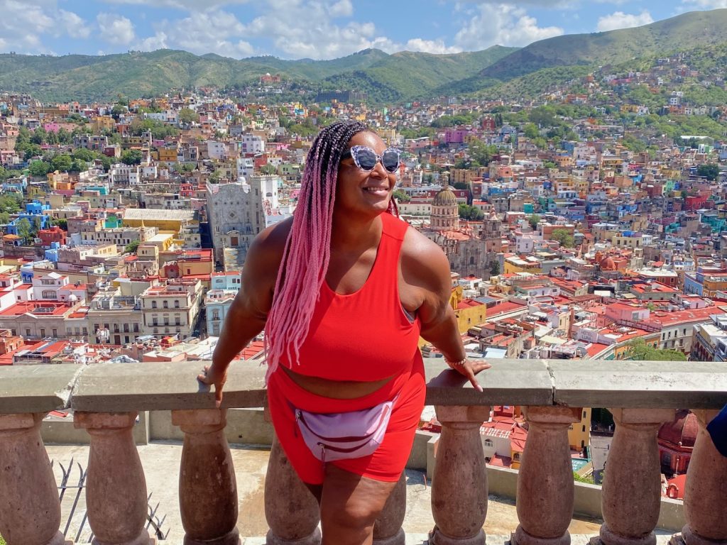 A woman wearing a red top and shorts leans against an ornate railing with a city backdrop