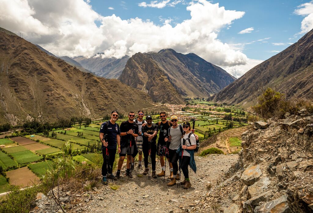 A group of travelers pose on a trek, a green valley and mountains behind them.