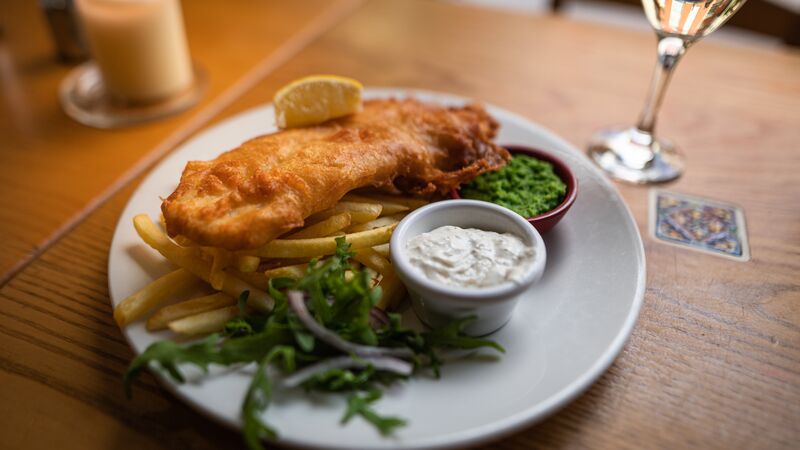 A plate of battered fish on a bed of crispy chips with a side of tartare sauce and mushy peas. 