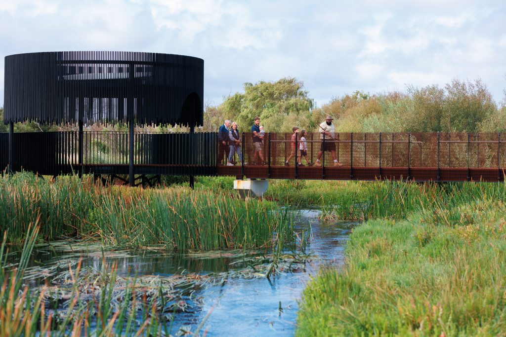 A group of people walking over a boardwalk crossing a marsh, with a modern black building behind them