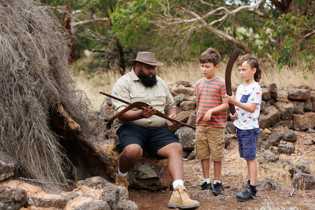 A man shows two children a selection of Indigenous weapons