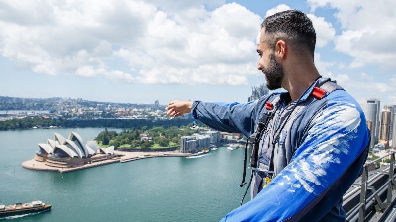 A man wearing a blue jumpsuit standing on top of a bridge, pointing to the Sydney Opera House