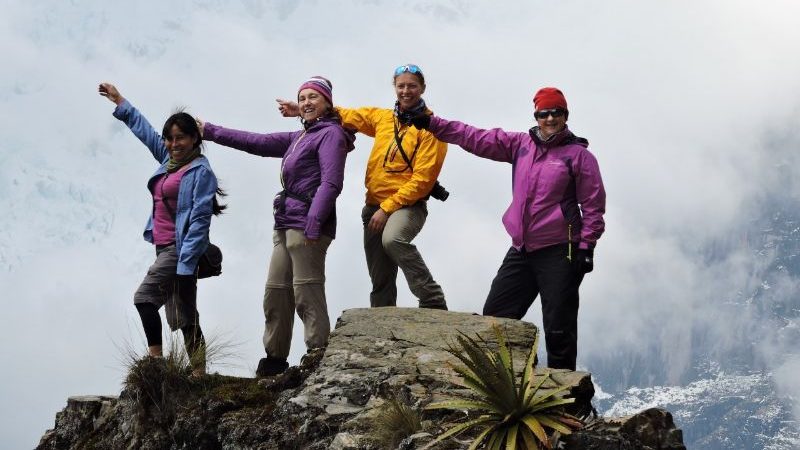 Four women in hiking gear standing on a mountain with their arms outstretched