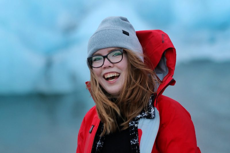 A young woman wearing a woollen hat, glasses and red jacket with icebergs in the background