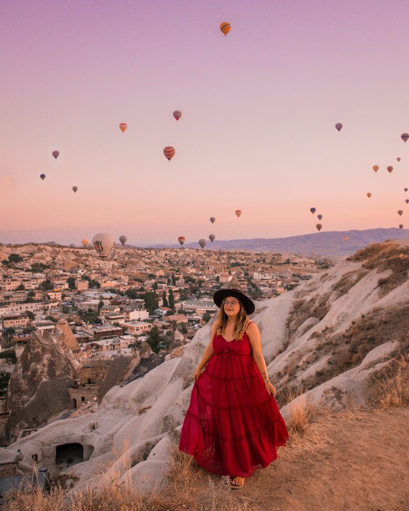 A woman in a red dress and hat admiring a dawn sky filled with hot air balloons