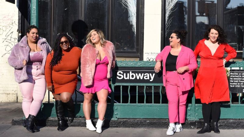 Five happy women in brightly coloured clothing standing next to a Subway sign