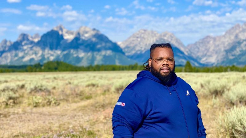 A man wearing a blue sports coat standing in a field with mountains in the distance